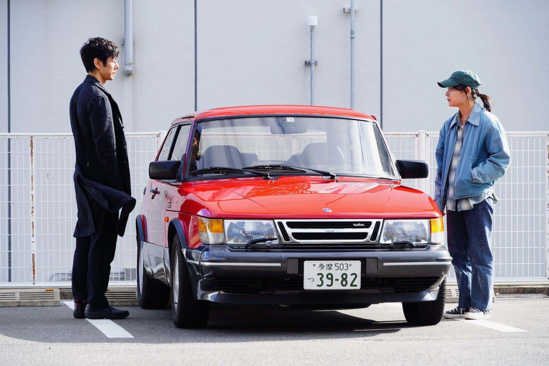 Promo still of two Drive My Car characters standing next to car