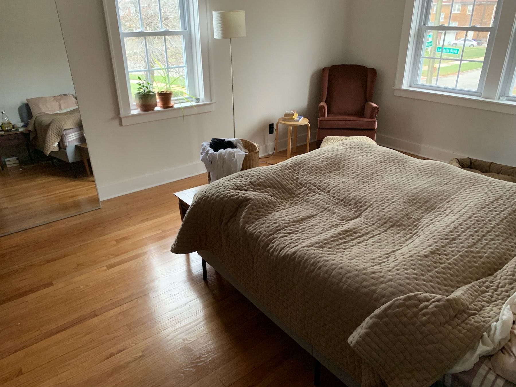 View of foot of the bed from the door next to the head of the bed. Windows on two sides of room, pink corner armchair, and tall mirror reflecting bed and side table.