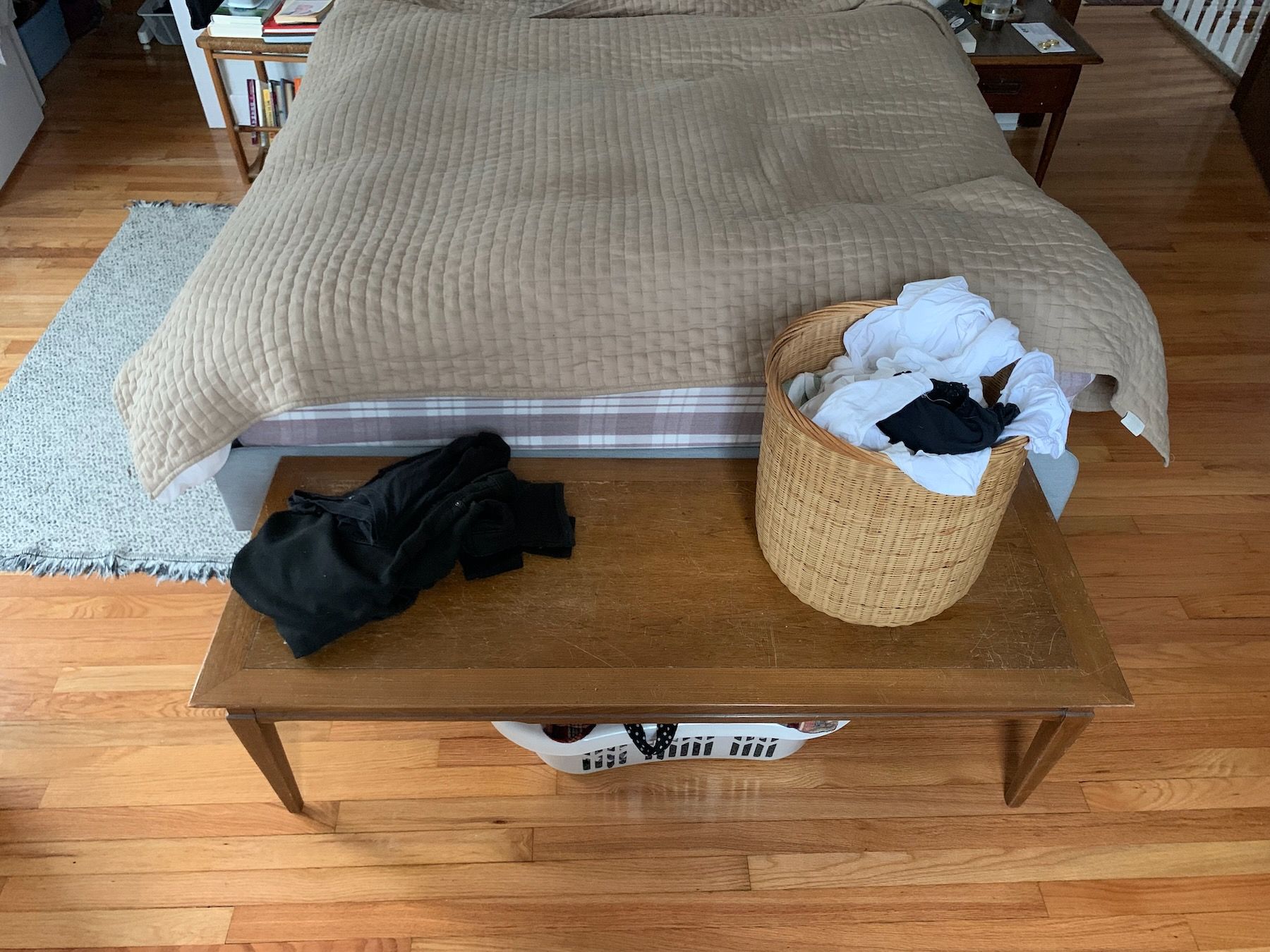 Close-up from a few feet away from foot of bed. Wooden coffee table bench with sharp corners, laundry baskets above and below, and small pile of black clothes.
