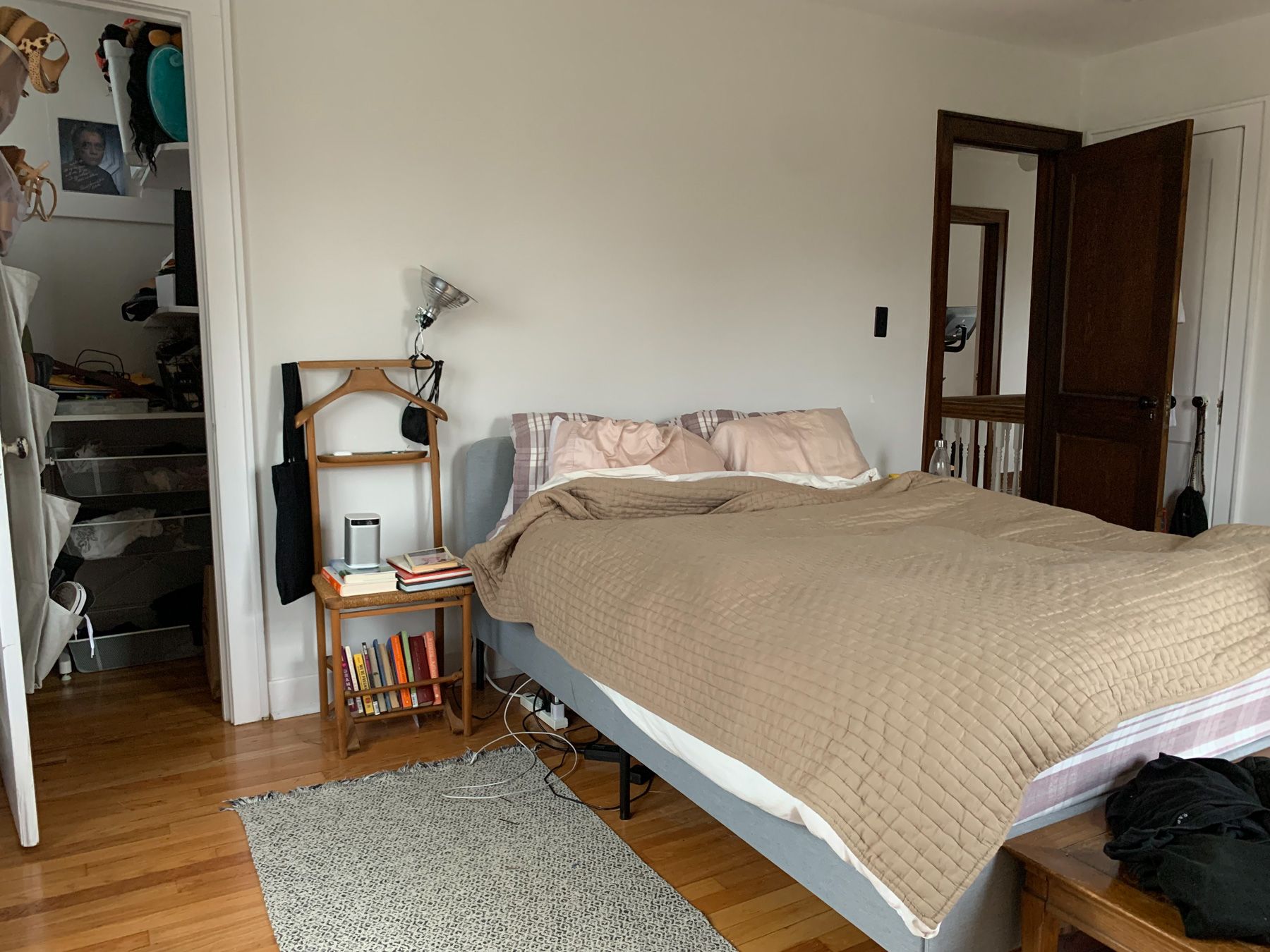 Similar view of bed from corner of room, except more centered at space around headboard. Wooden Italian butler chair with books on it, used as a side table. Clip lamp aimed at blank wall above headboard.