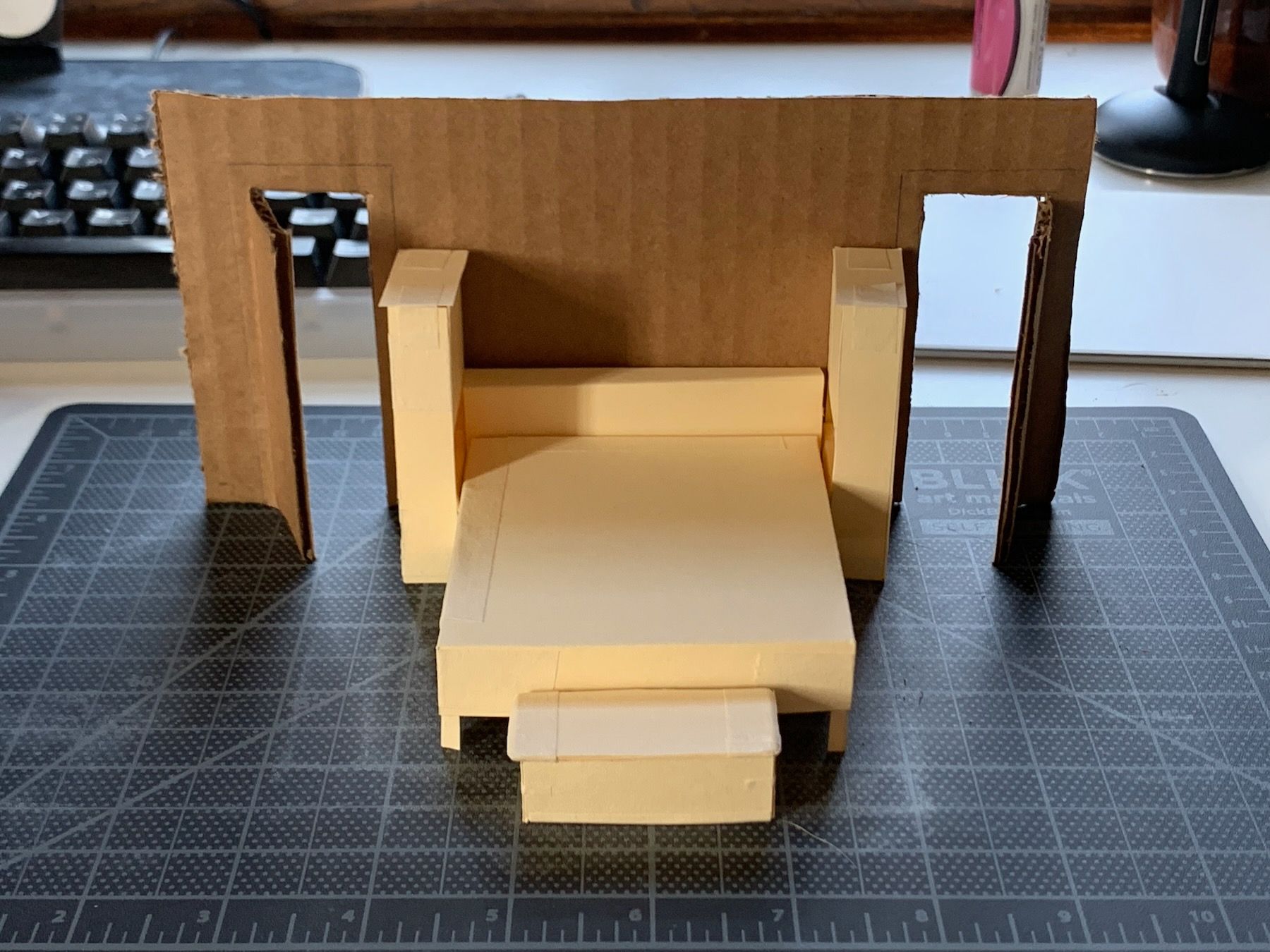 Scale model, furniture made of manilla folder stock and back wall and doors made of cardboard. Standing on a black cutting mat on desk, keyboard and mousepad in background. Elevation view toward wall from beyond foot of bed.