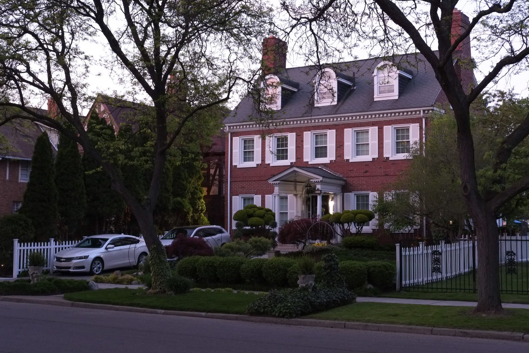 Dusk light, stately brick house with white shutters, well-appointed front yard shrubs.