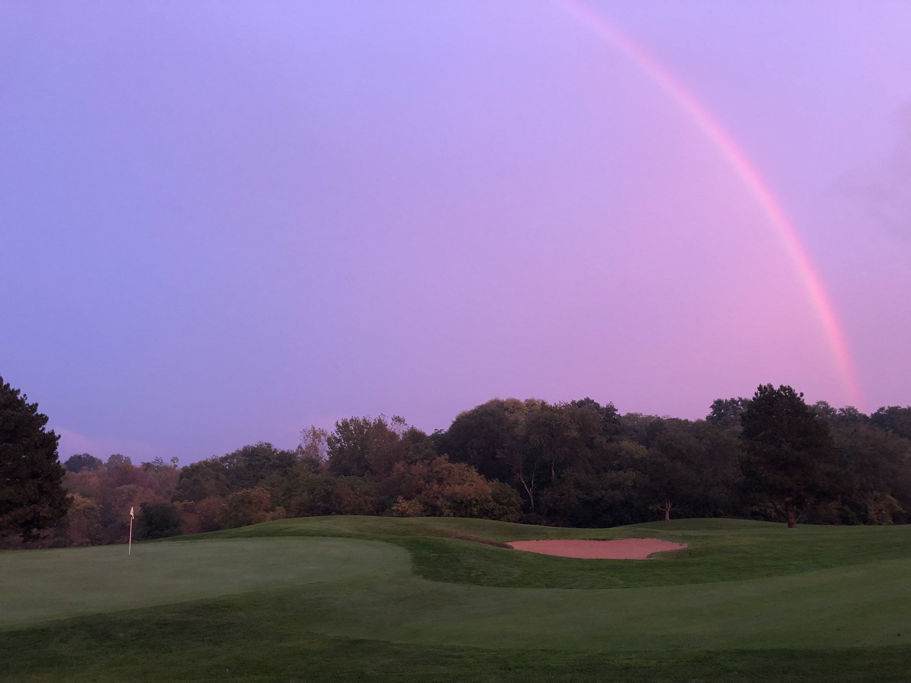 Purple-skied rainbow over a golf course. Rolling hills, green with flag in lower left corner.