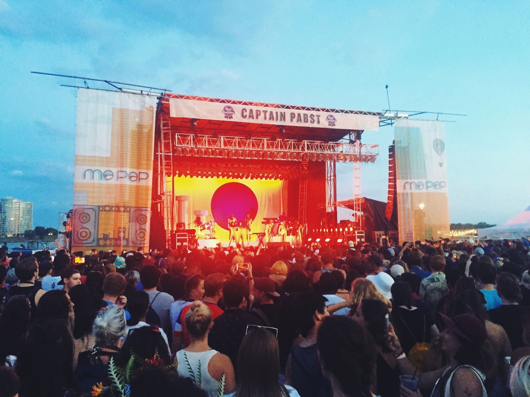 Orange lit stage at dusk. Crowds of people.