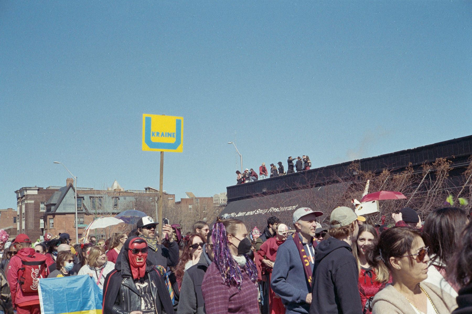 Marche du Nain Rouge marchers, Ukranian flag and sign, watchers on rooftop in background, clear blue sky.