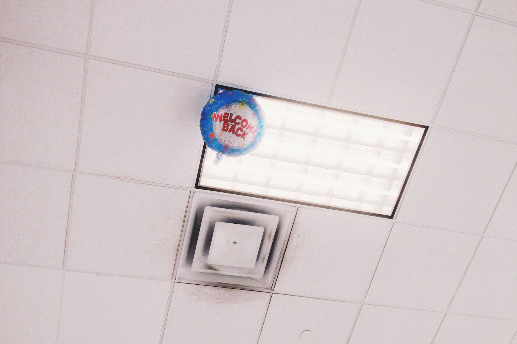 Black and white. Escaped 'Welcome Back' balloon against drop ceiling fluorescent light.