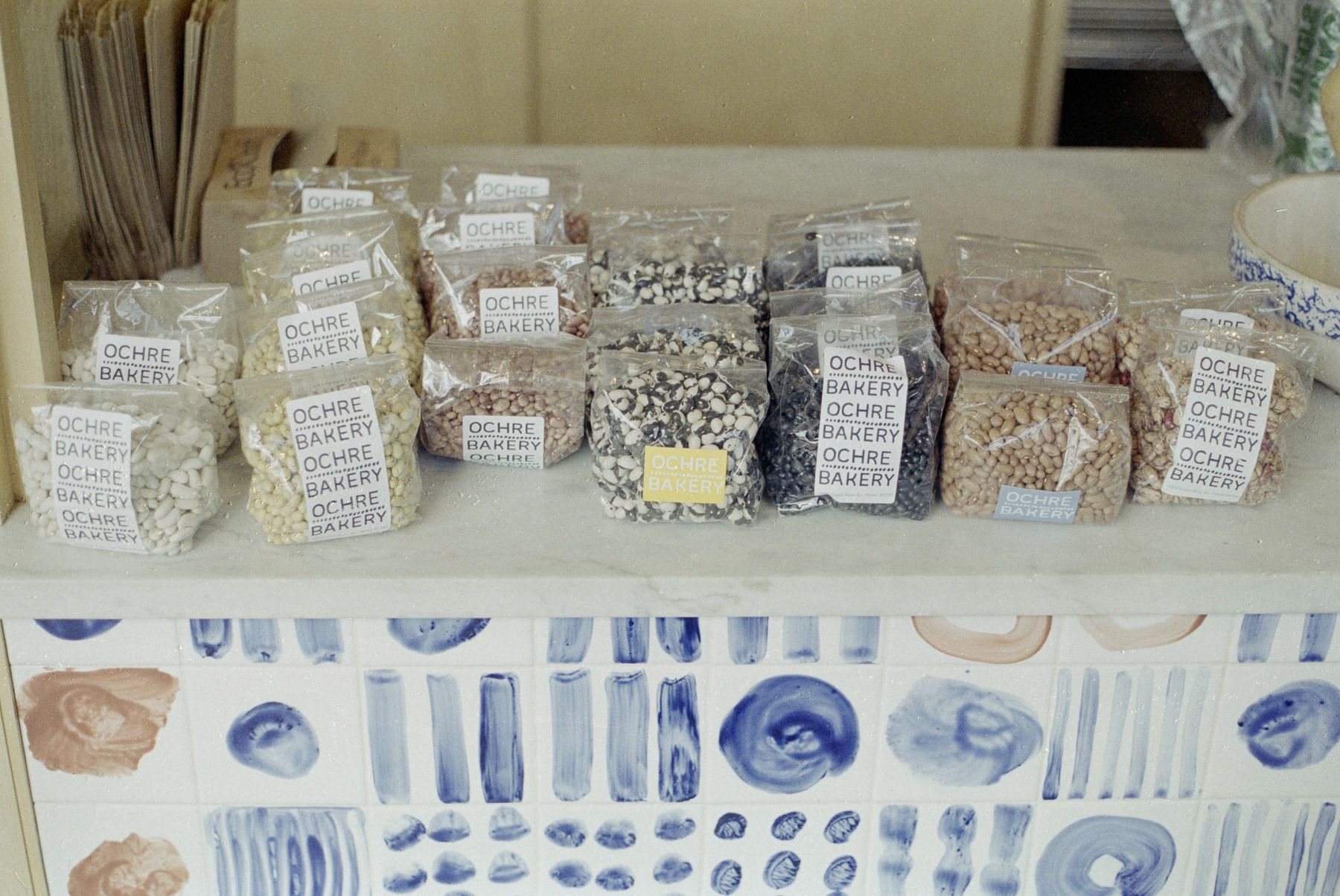Dried beans in clear bags labeled Ochre Bakery, on marble counter with hand-painted tile front.