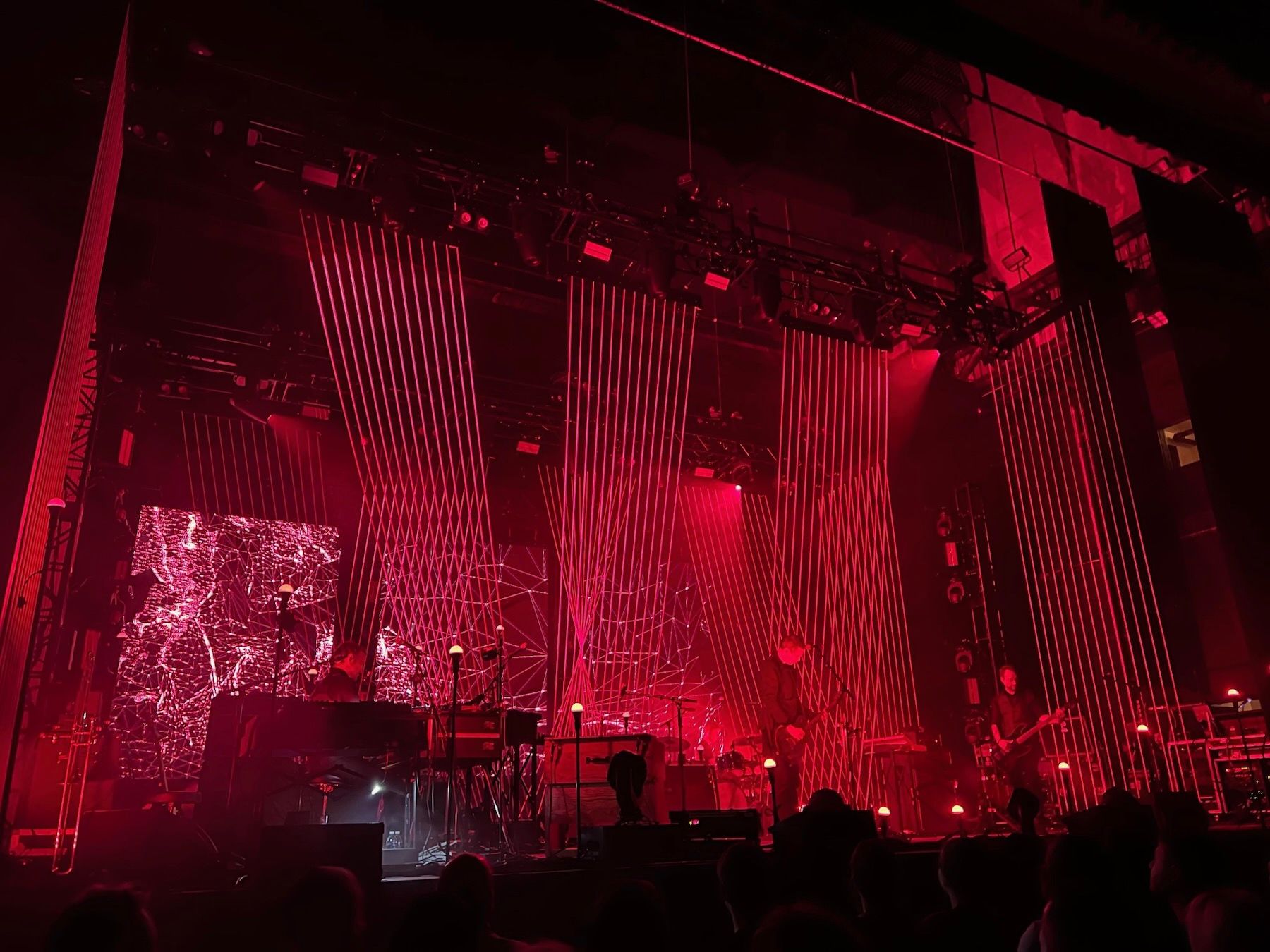 Moody red-lit stage with band, set decorated with groups of thin parallel rods running from stage to ceiling.