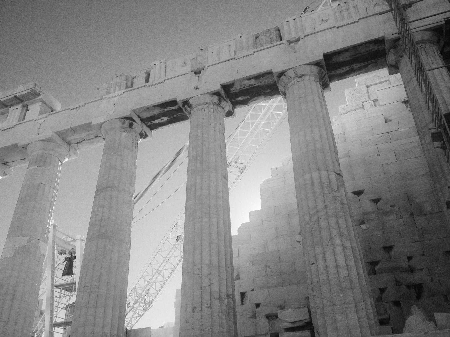 Black and white. Doric columns at the Parthenon, metal crane in background with glow of sun.