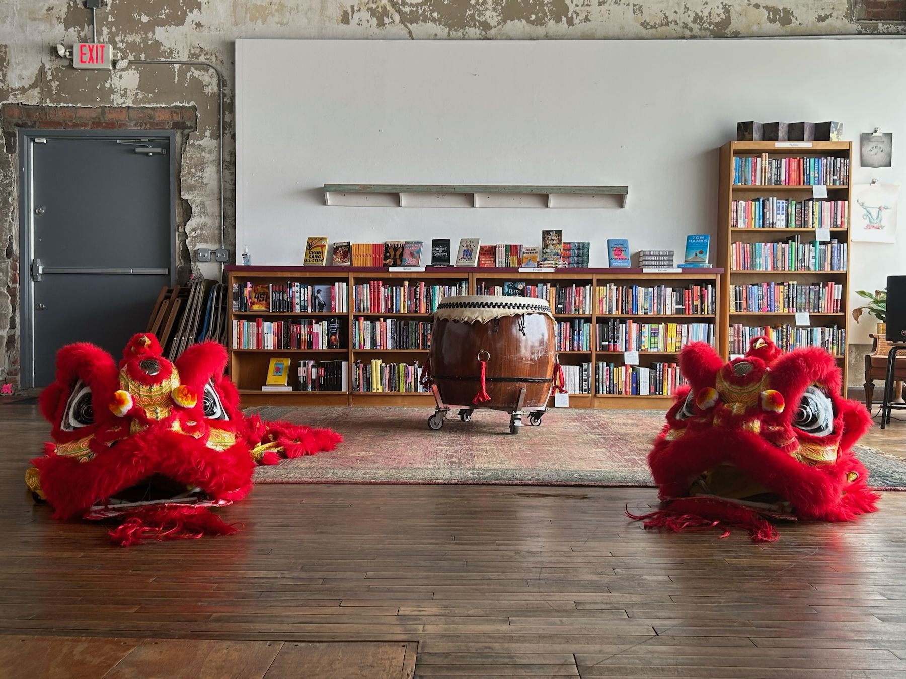 Two red lion mask heads and barrel drum awaiting performance in front of bookshelves.