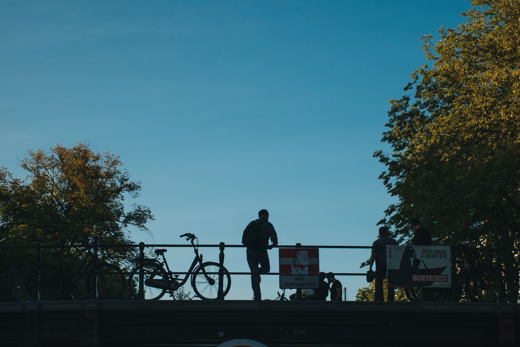 Silhouette of man on canal bridge looking at phone, bicycle next to hi