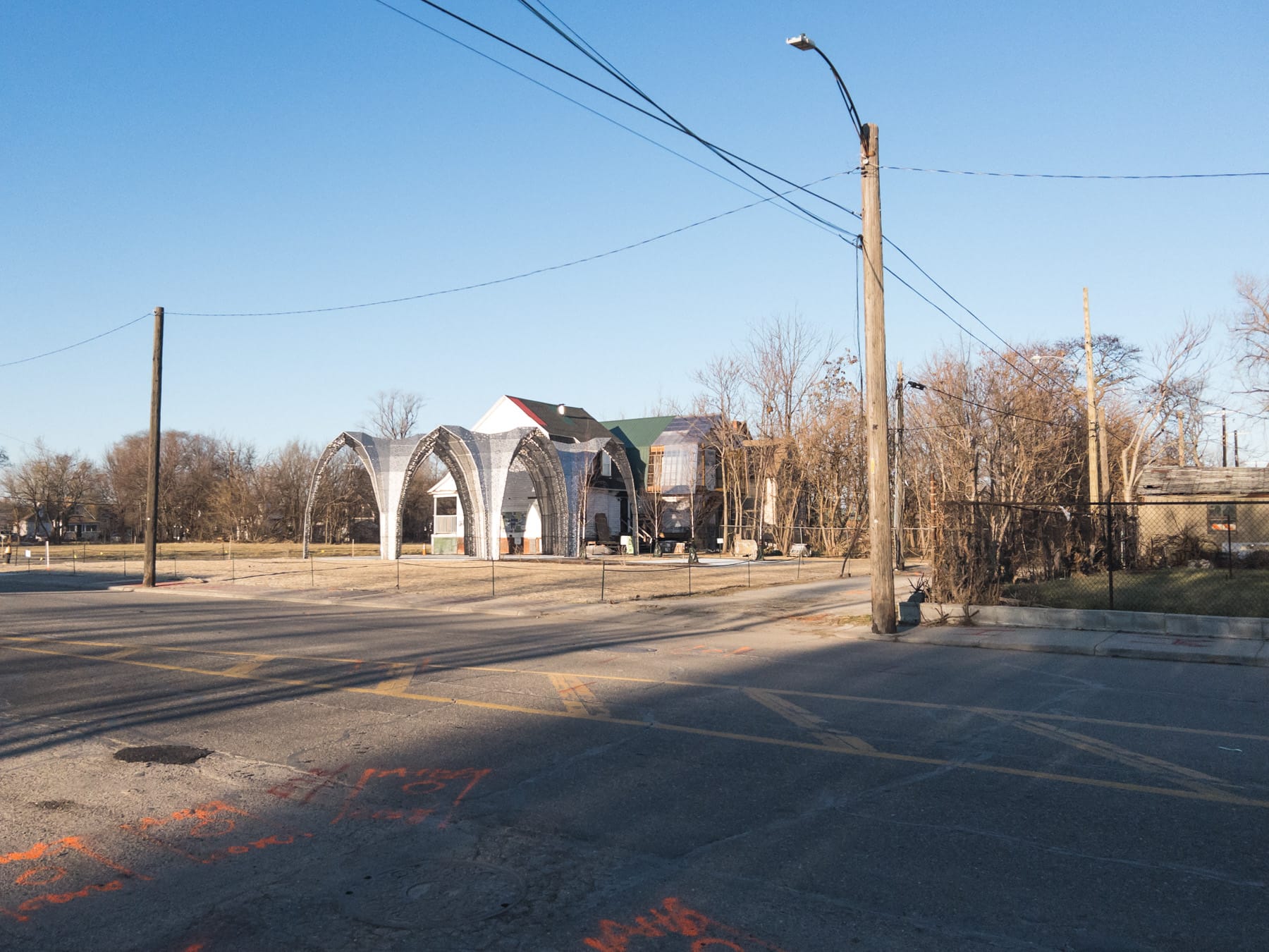 Wide city street with faded lane lines and utility spray. Laser cut metal pavilion in front of under-construction house.