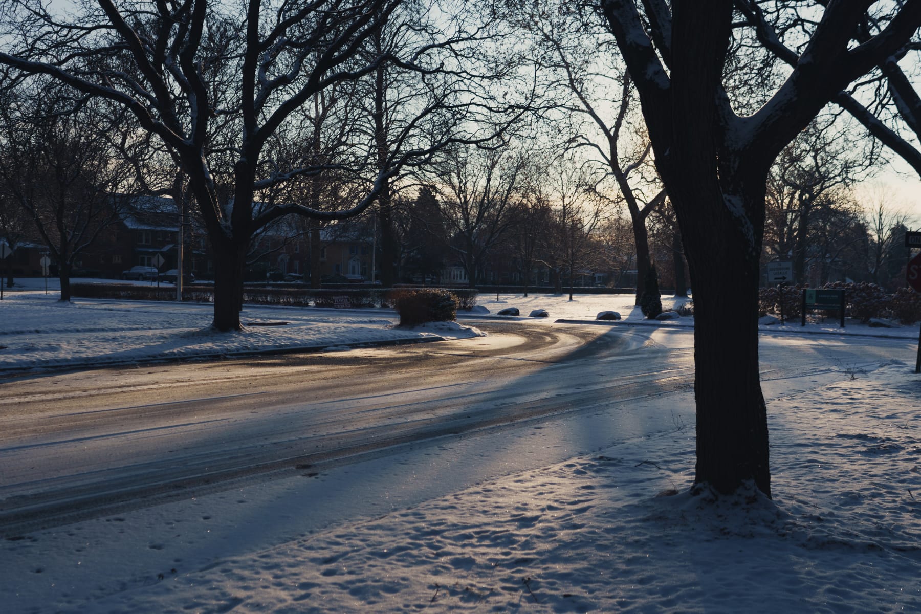 Snowy park street corner in low morning light, streaks of tire tracks, ice crystals aglow.