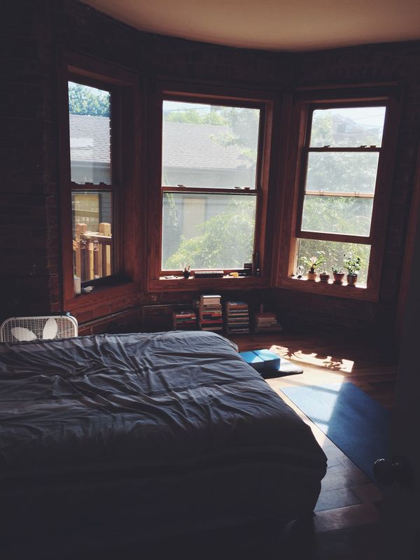 Sparse, sun-lit bedroom. Yoga mat on floor, box fan, stacks of books, plants in window.