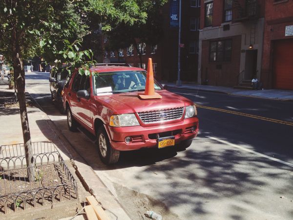 Orange traffic cone on the hood of a red SUV; an urban unicorn. 