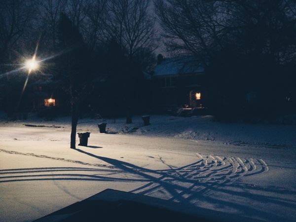 Snowy street at night, tire tracks carved into driveway. Streetlight glare, a couple of porch lights and garbage cans.