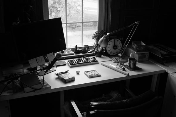 Black and white. White desk with computer monitor next to window, looking out onto trees. Clay, tile, tools, keyboard.