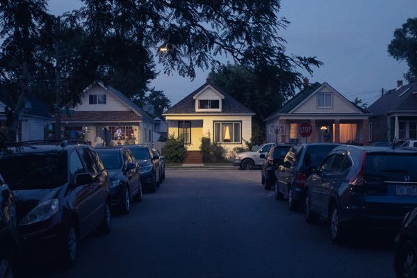 Dusky evening. Narrow car-lined street at three small bungalows, middle one lit warm yellow by streetlight.